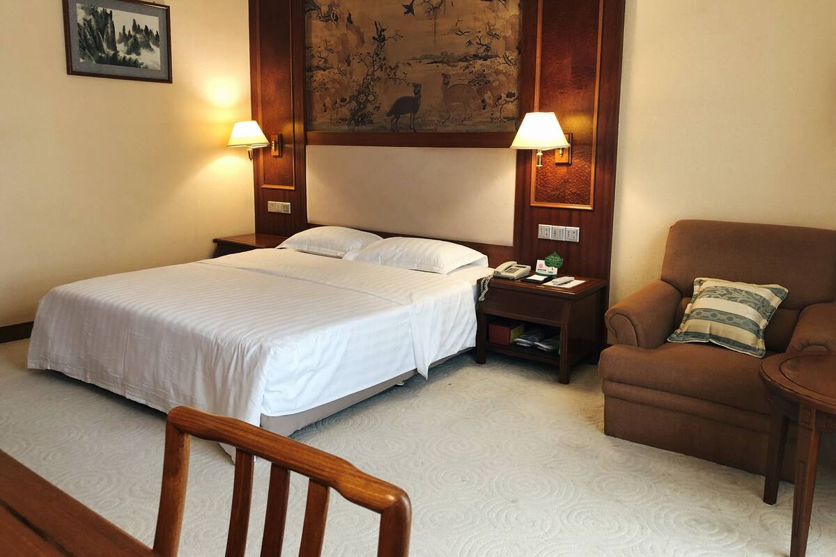 A hotel room with a large bed, two bedside lamps, a wooden headboard, a chair, and a small table.