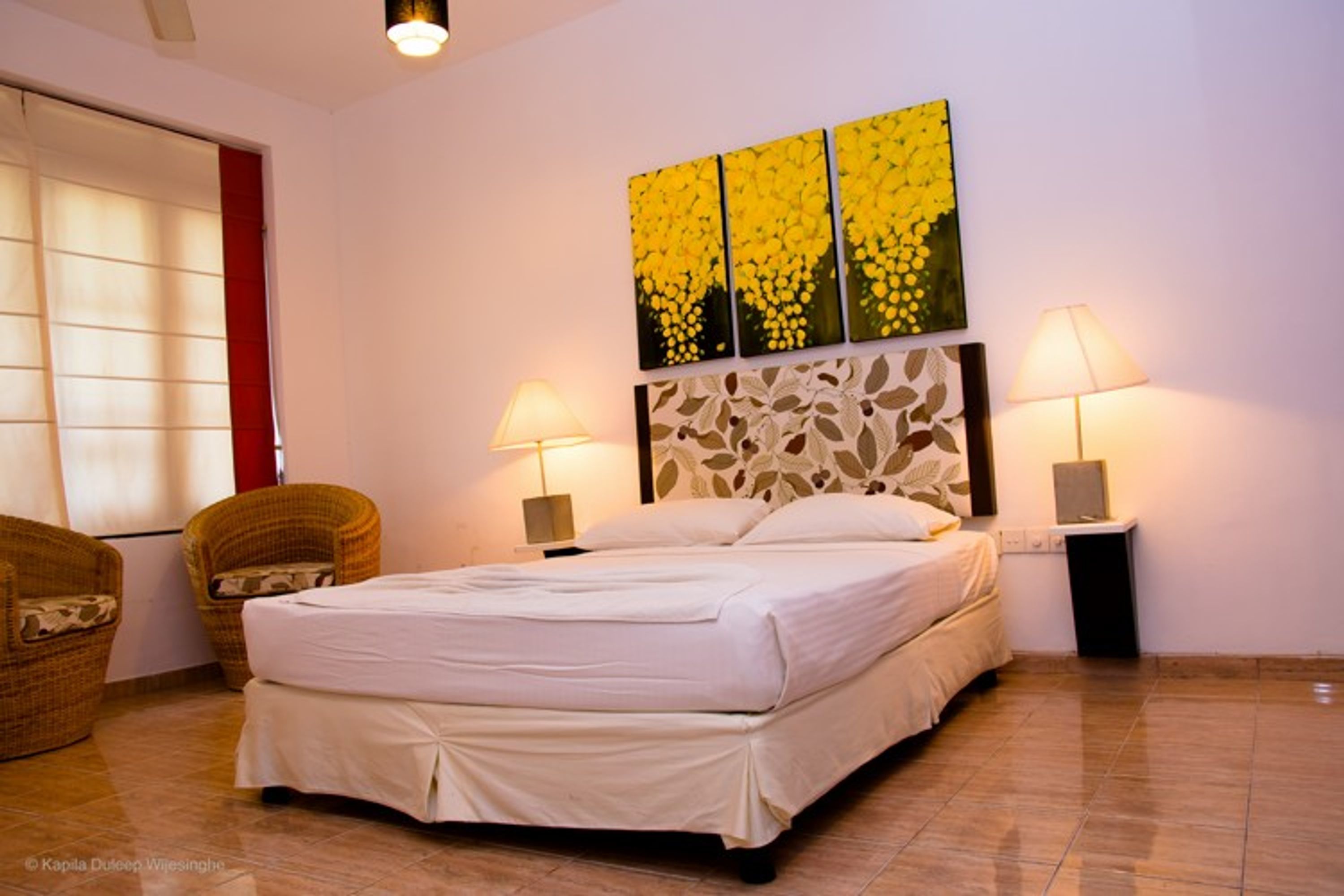 Quarto com cama, duas mesas de cabeceira, duas luminárias fixadas na parede, uma obra de arte na parede e duas cadeiras de vime.