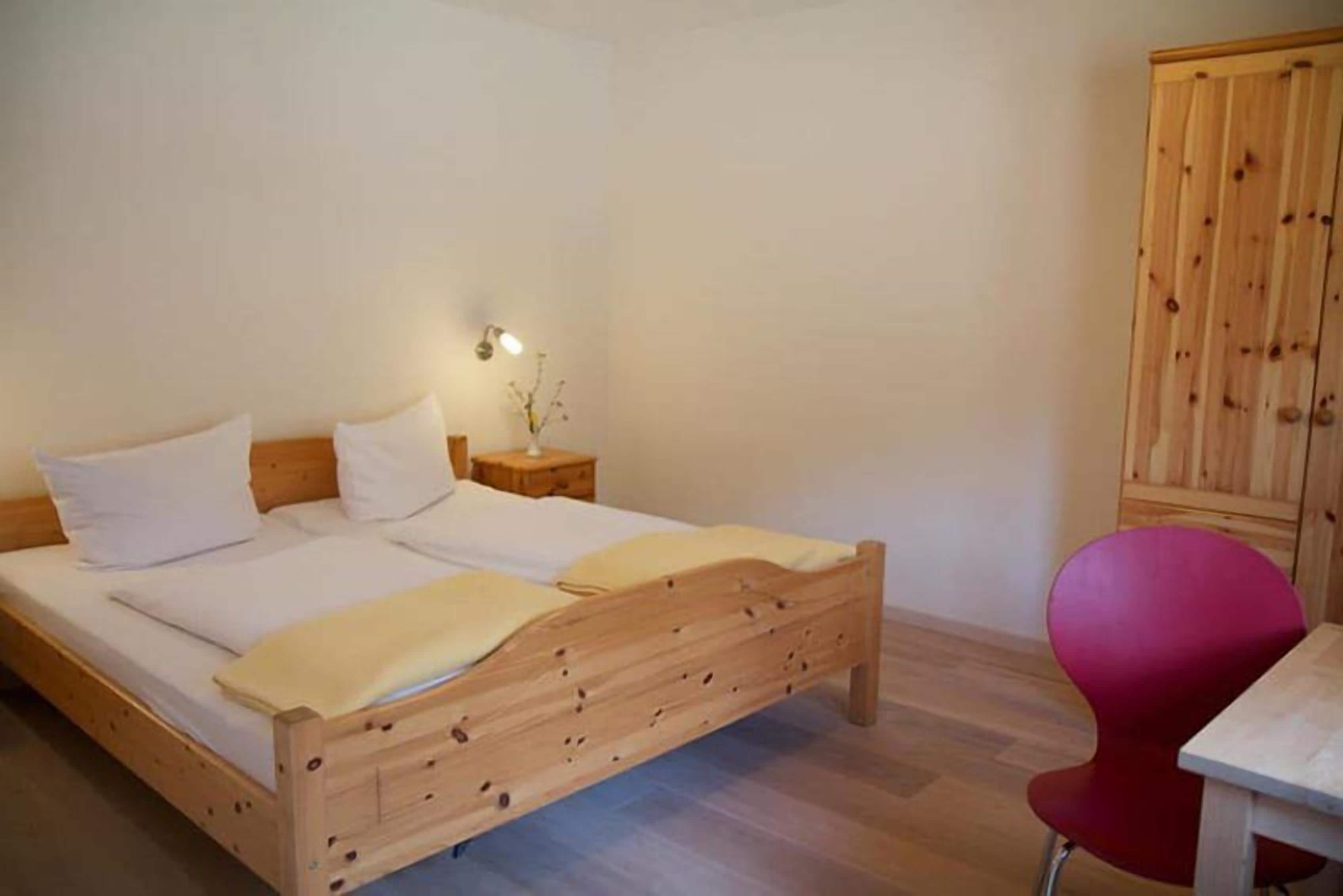 A wooden bed with white bedding, a nightstand with a lamp, and a red chair in a simple room with wooden flooring and a wardrobe.