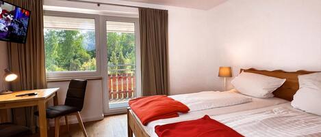 Habitación de hotel con cama, escritorio, silla, televisor y balcón con cortinas.