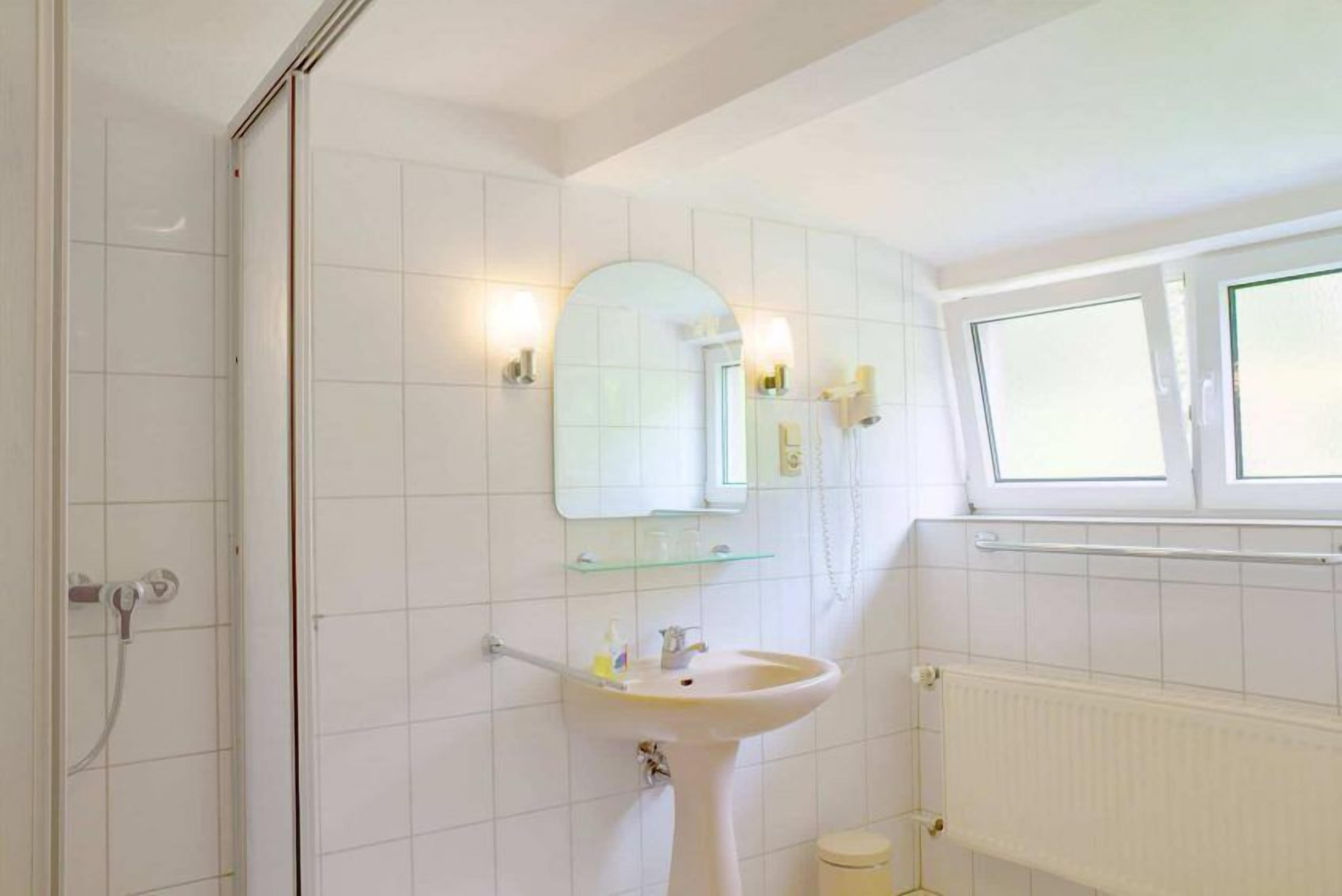 A bathroom with white tiles, a pedestal sink, a mirror with lights, and a window.