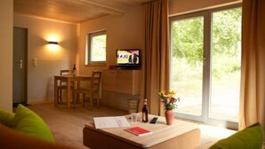 A cozy room with a wooden ceiling, a dining area with a table and chairs, a TV on the wall, and a large window with curtains. - Biohotel Spöktal (Bispingen)