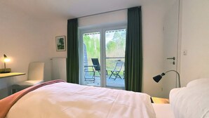 A hotel room with a bed, a desk with a lamp, a chair, and a view of an outdoor deck with chairs. - Biohotel Spöktal (Bispingen)