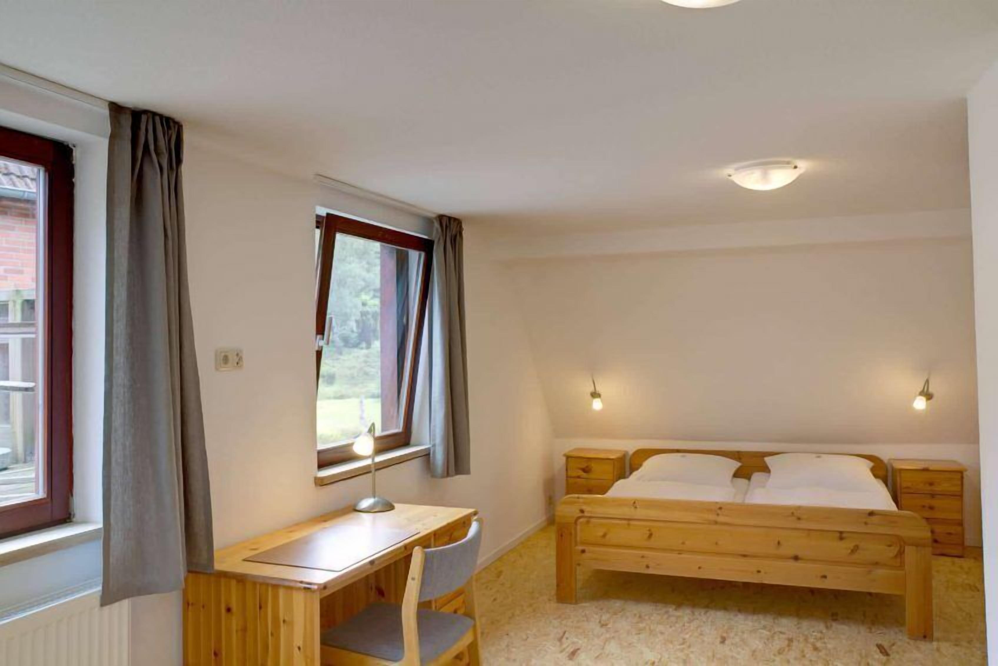 A bedroom with a wooden bed, a desk with a chair, and a window with curtains.