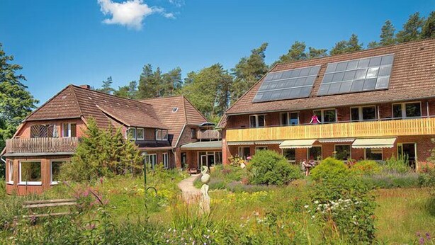 A rustic wooden building with a balcony and a second-story balcony, surrounded by lush greenery and a clear sky. - Biohotel Spöktal (Bispingen)