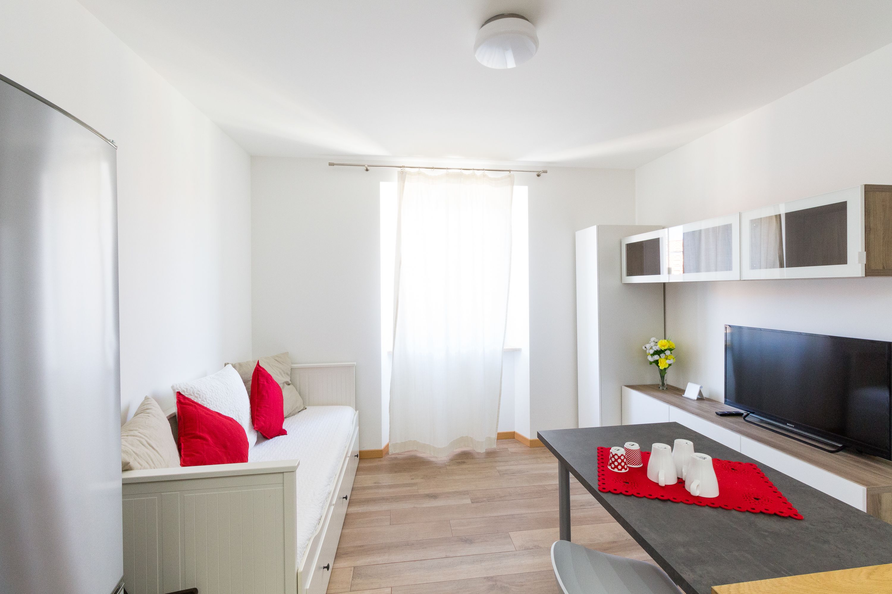 Quarto moderno e bem iluminado, com sofá, mesa de jantar com jogo americano vermelho, televisão de tela plana e janela com cortinas leves.