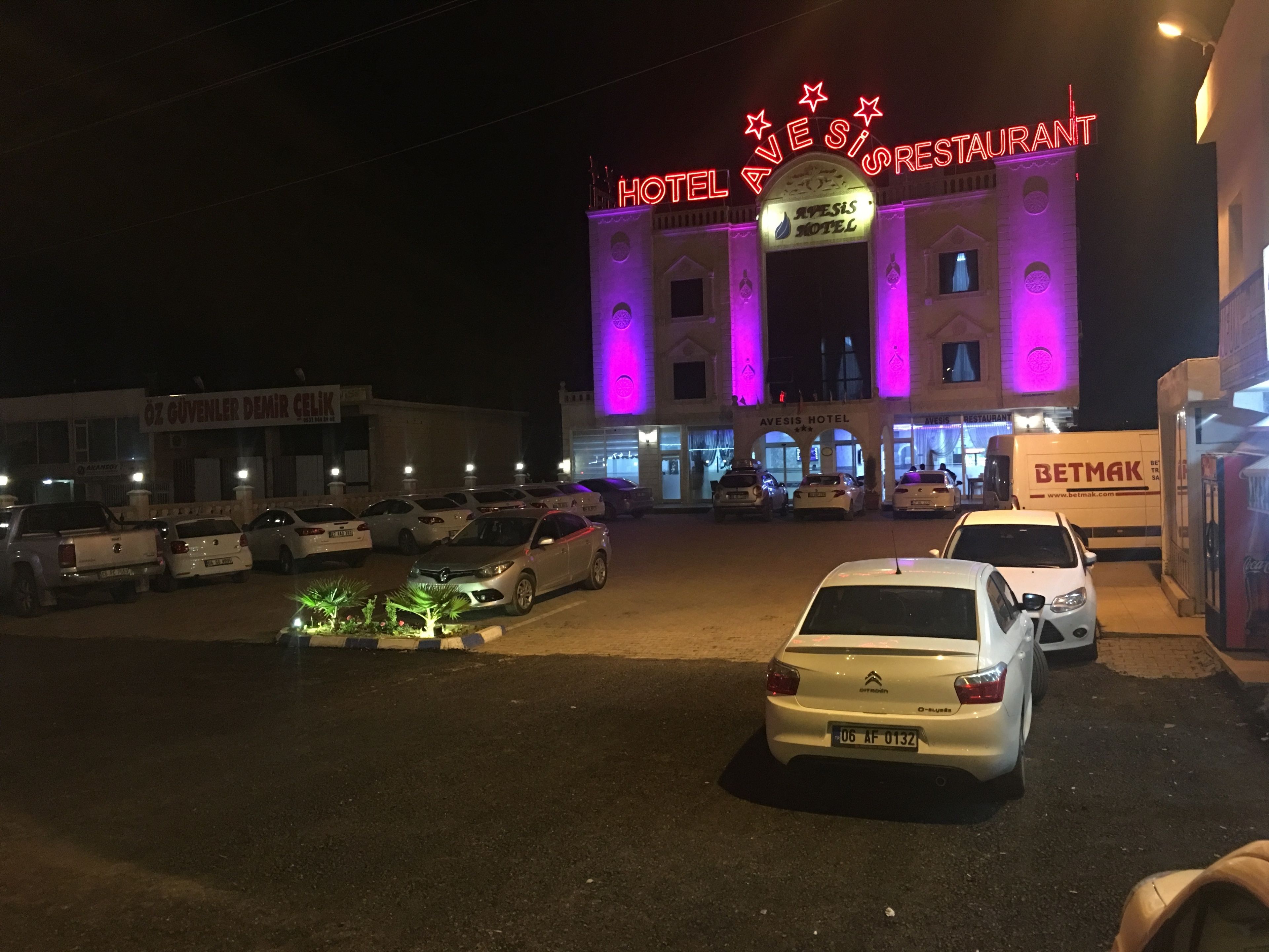 A hotel with a neon sign, multiple parked cars, and a Betmak delivery truck.