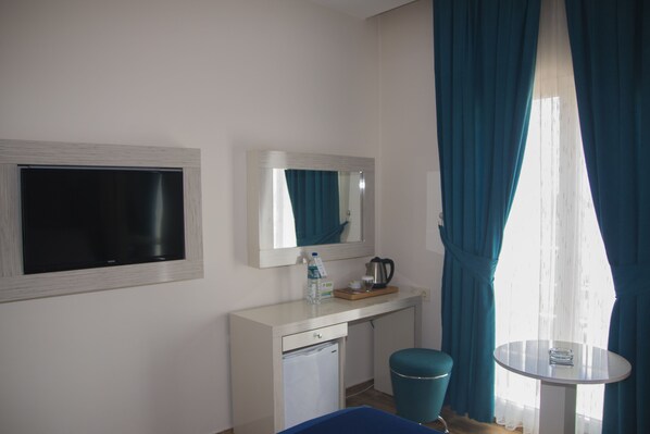 A hotel room with a TV, a mirror, a small table with a cup and a kettle, a water bottle, and a blue chair. - Avesis Hotel (Nusaybin)