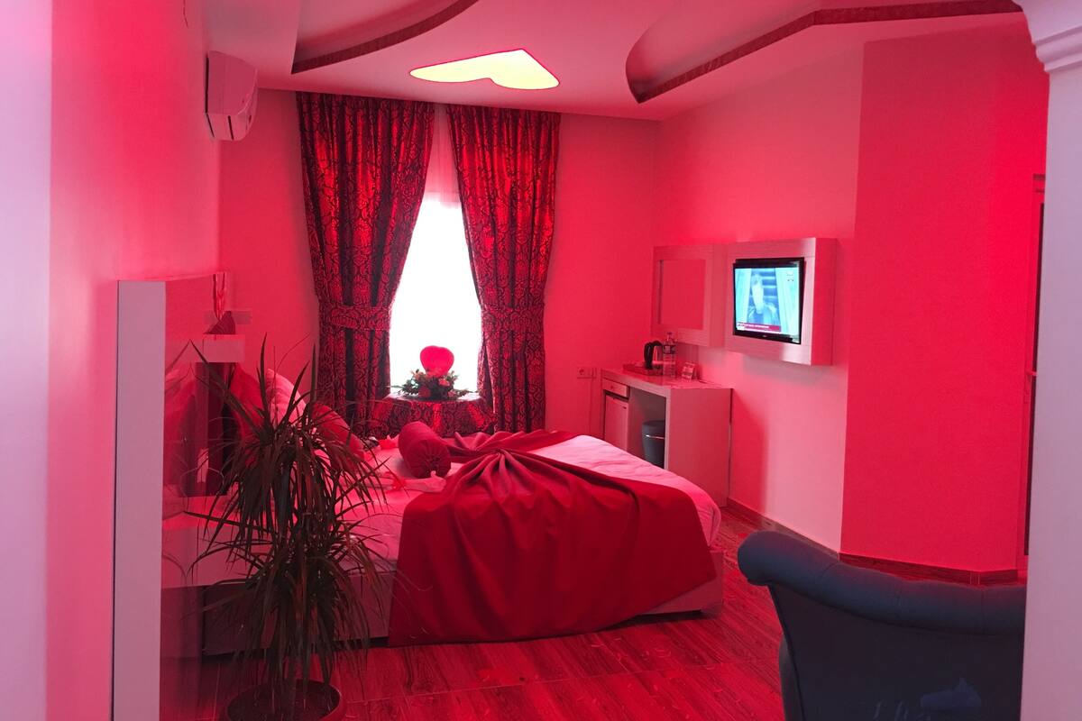 A bedroom with a red bed, a television on the wall, and a window with red curtains.
