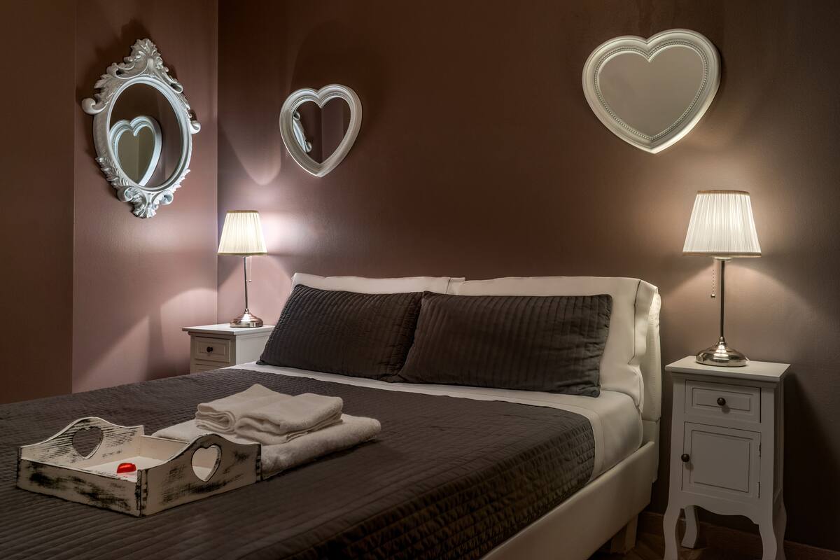 Un letto rifatto con cura, con una coperta scura, due abat-jour, uno specchio a forma di cuore e un comodino con una lampada.