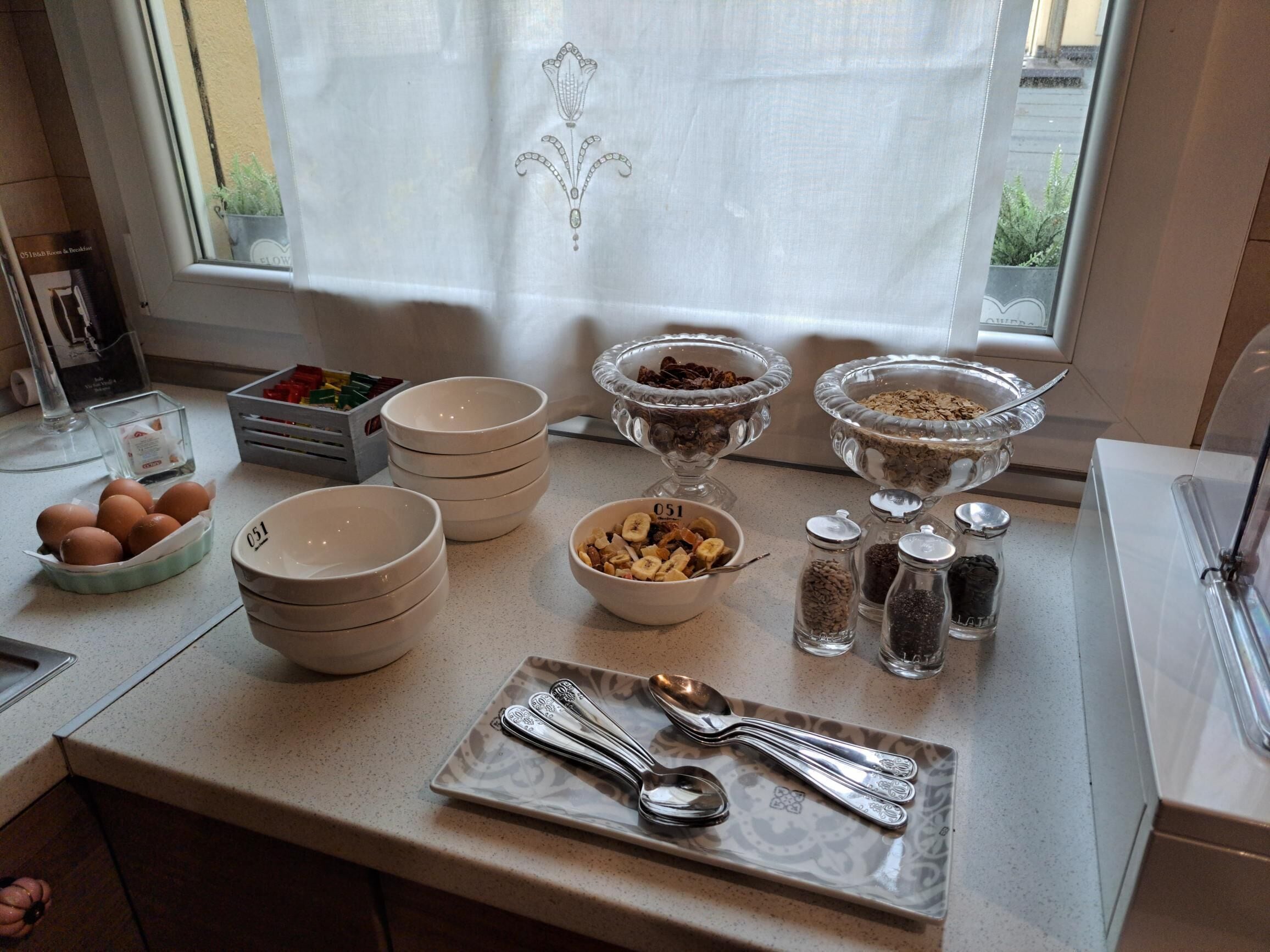 En köksbänk med olika skålar som innehåller matvaror, en tallrik med bestick och ett fönster med en gardin.