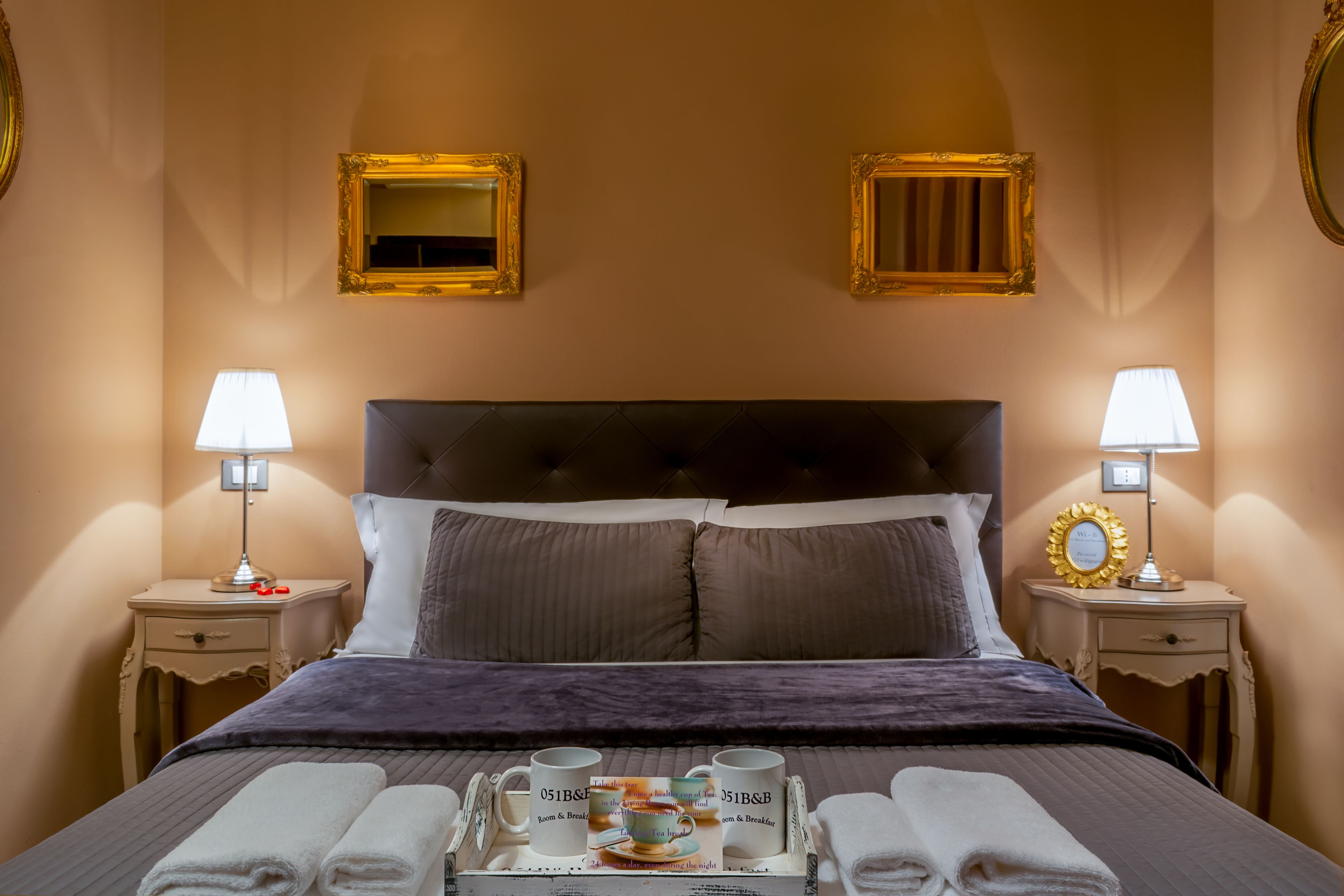 A neatly made bed with a tray of breakfast items, two bedside lamps, and framed mirrors on the walls.