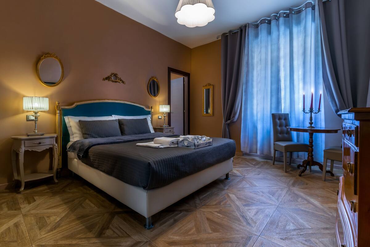 Una camera d'albergo con un letto grande, due comodini con lampade, una cassettiera, una sedia e un tavolo con un candelabro.