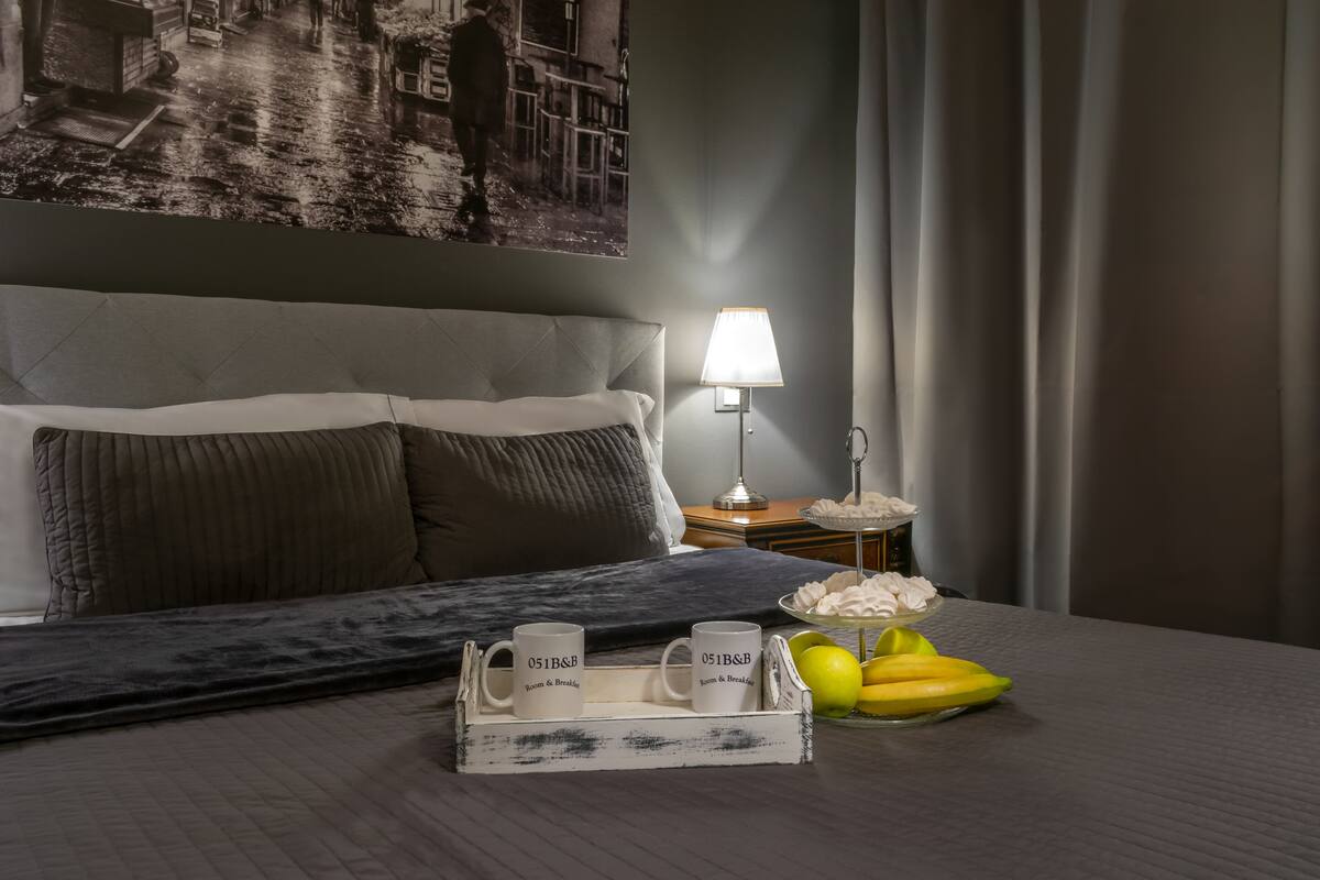 Un letto rifatto con cura, con una testiera grigia, un comodino con una lampada e un vassoio con due tazze e una ciotola di frutta.