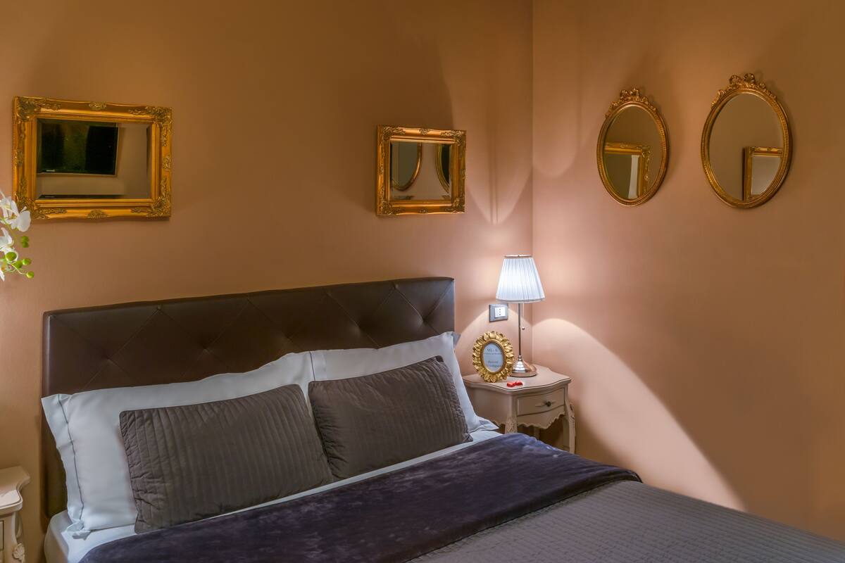 Una camera da letto con un letto grande, due specchi fissati al muro, un comodino con una lampada e un piccolo oggetto decorativo.
