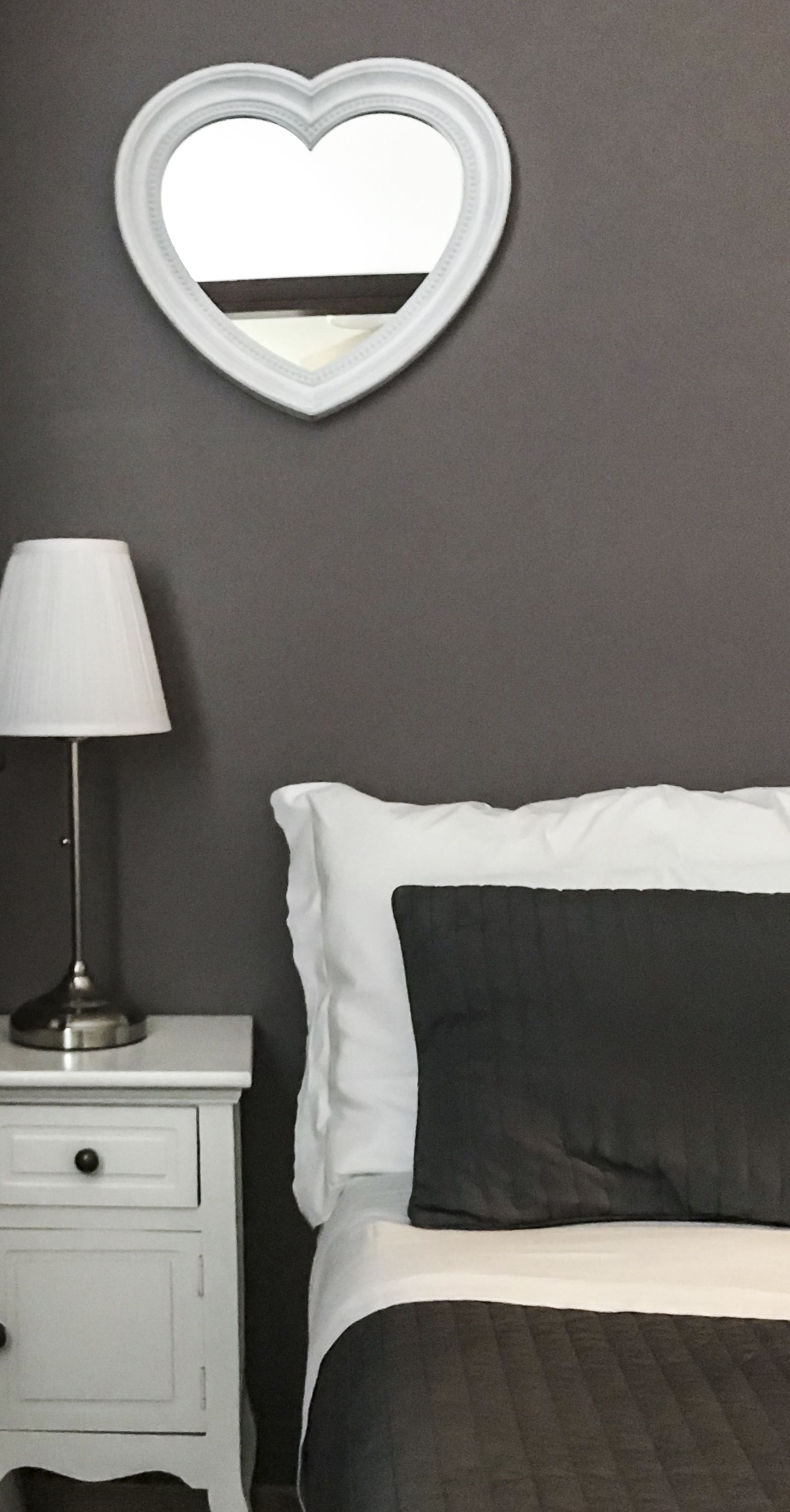 A bedroom with a bed, a nightstand, a lamp, and a heart-shaped mirror on the wall.