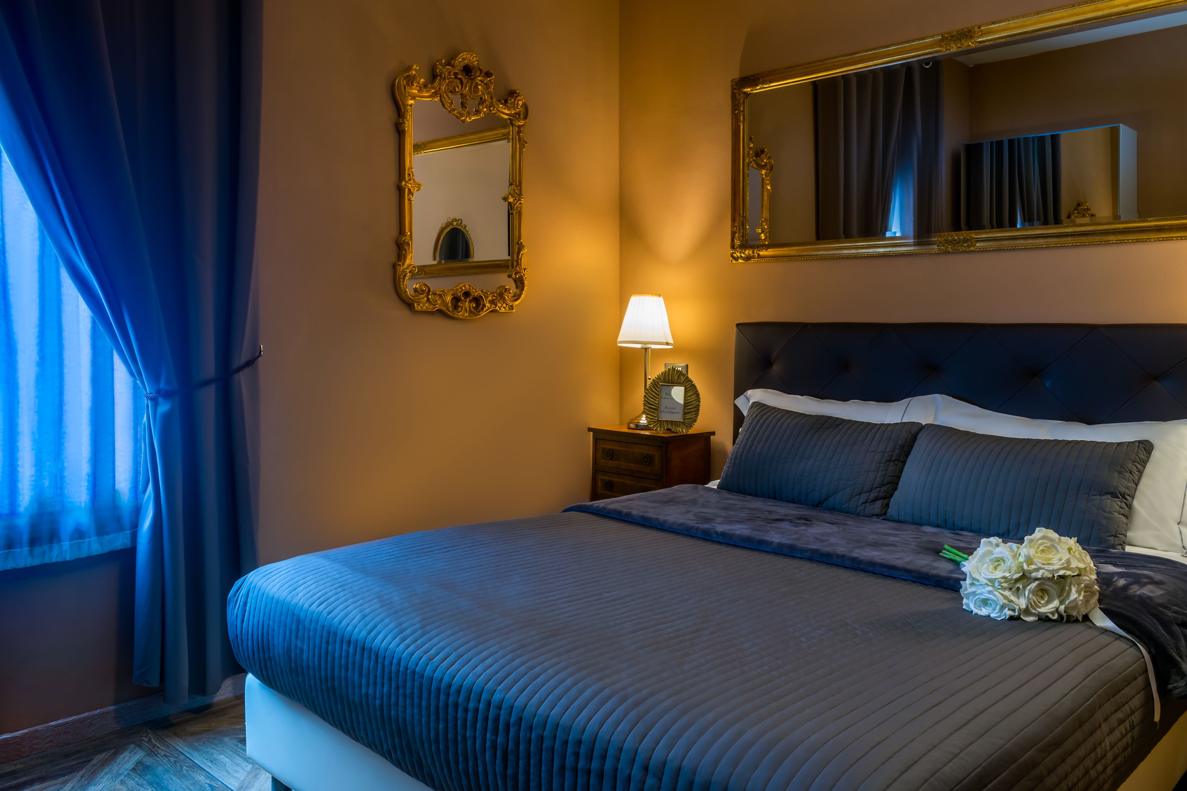 A hotel room with a large bed, a nightstand with a lamp, a mirror, and a window with curtains.