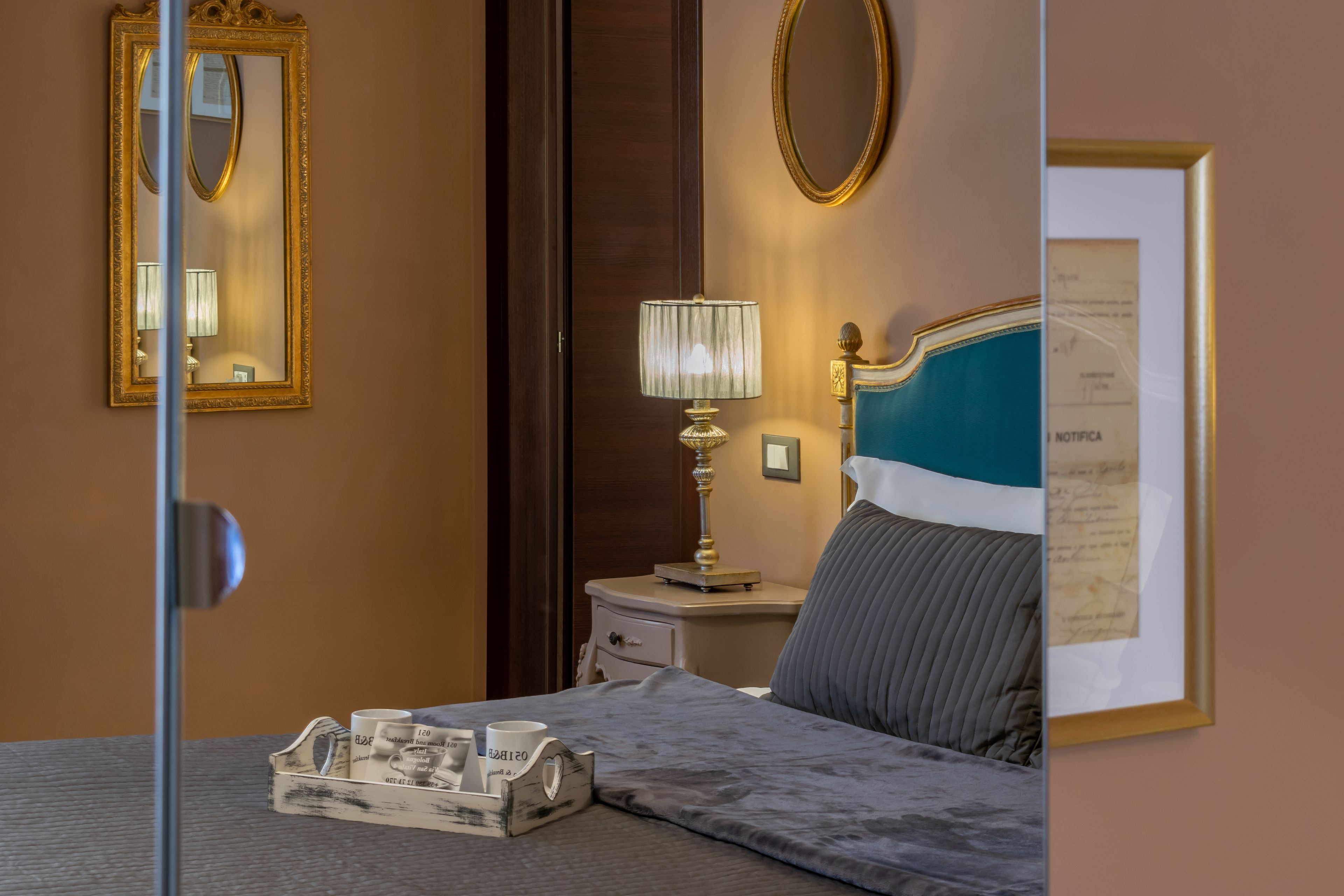 Una habitación de hotel con una cama, una mesita de noche, una lámpara, un certificado enmarcado en la pared y un espejo.