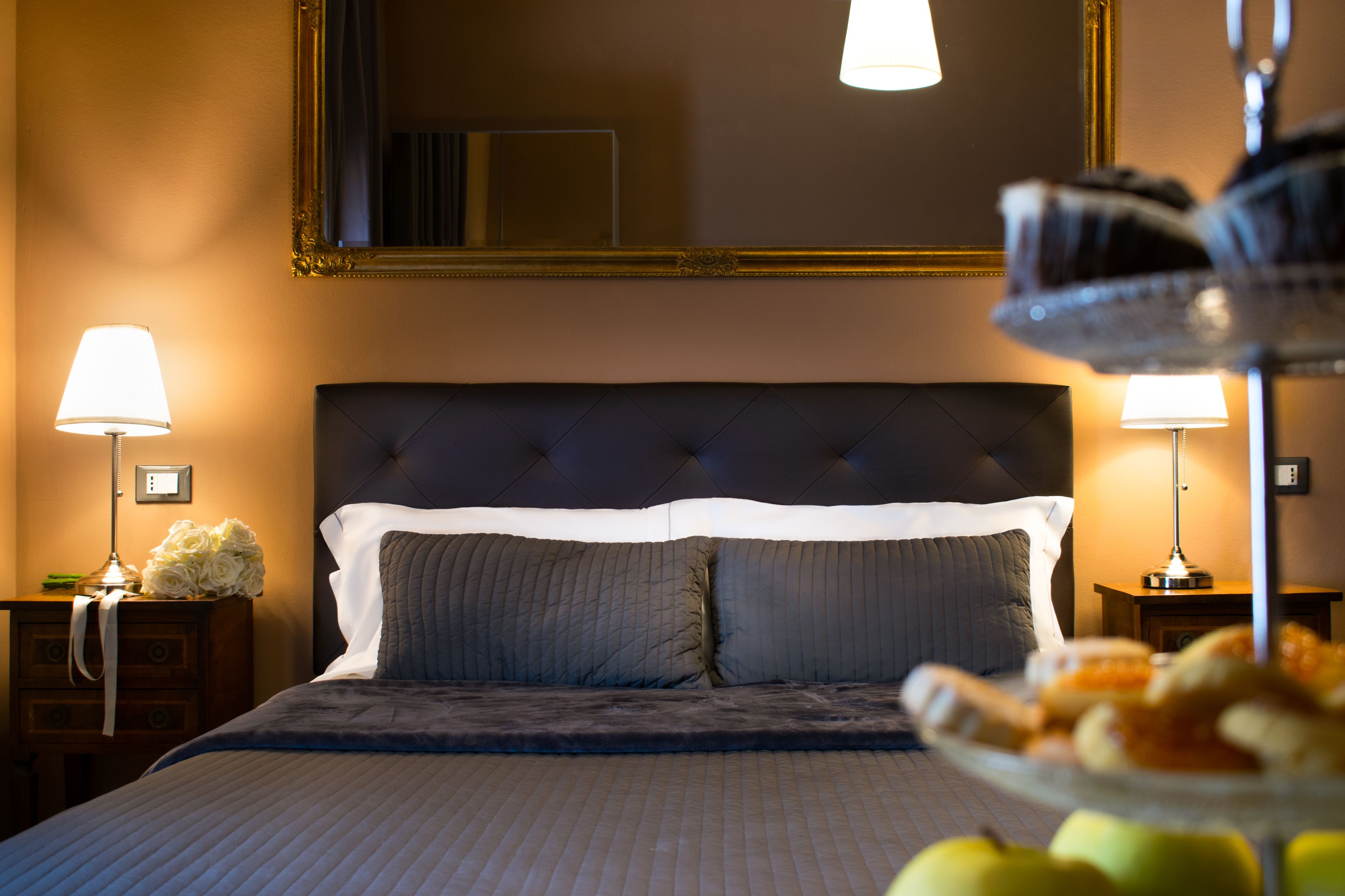 Een opgemaakt bed met een witte deken en grijze kussens, met aan beide kanten nachtlampjes op houten nachtkastjes.