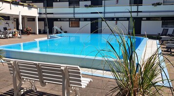 Uma piscina retangular com superfície de água azul cristalina, ladeada por um deck revestido com azulejos. Há espreguiçadeiras e um banco ao longo da borda da piscina.