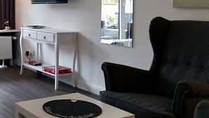 A modern living room with a grey sofa, a white side table, a flat-screen TV mounted on the wall, and a window with a view of the outside. - Apartment Regina am schönen Möhnesee (Moehnesee)
