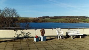 A waterfront terrace with a bench and chairs, overlooking a lake and distant hills. - Apartment Regina am schönen Möhnesee (Moehnesee)