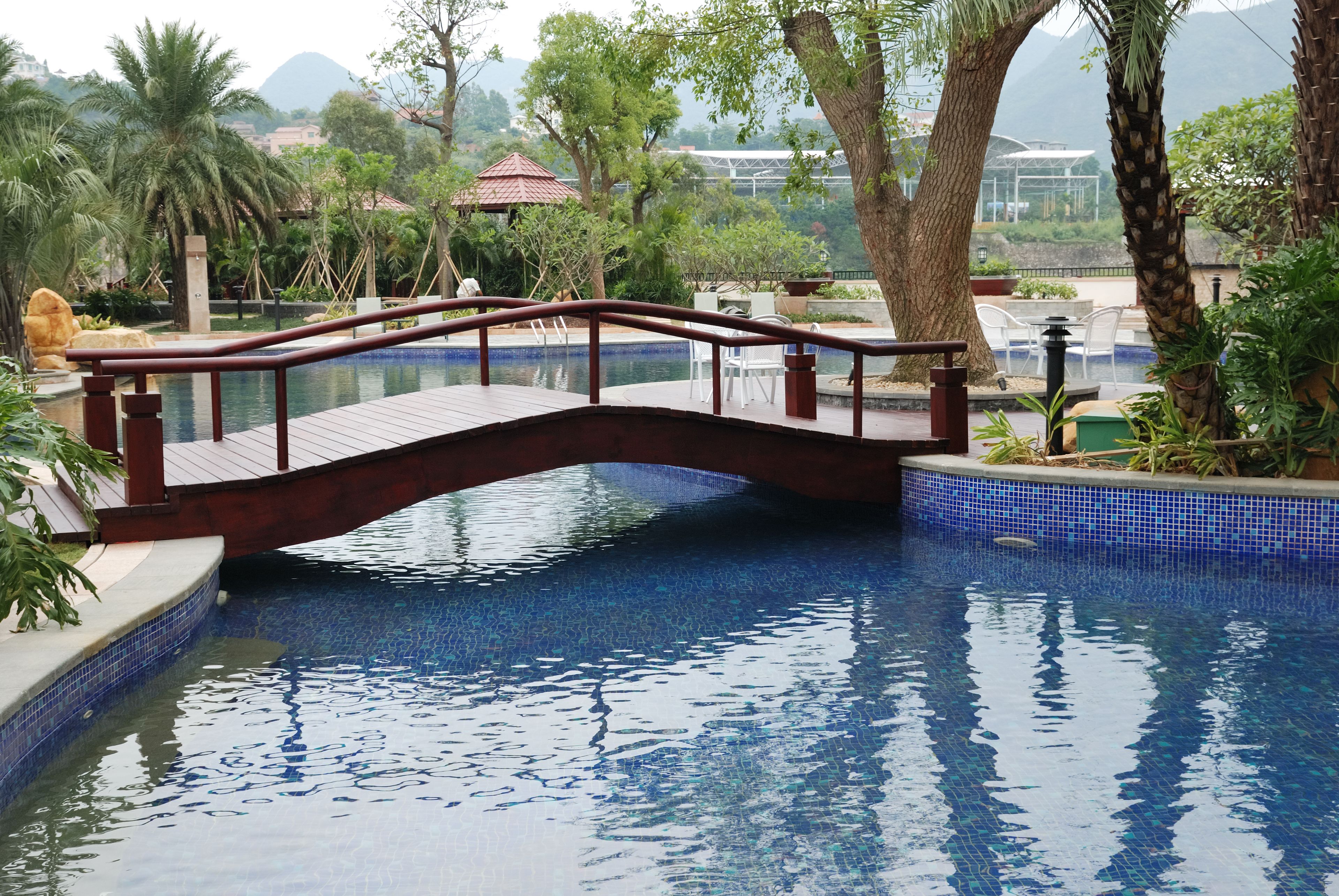 Un puente de madera sobre una piscina azul cristalina con bordes de azulejos, rodeado de exuberante follaje y árboles.
