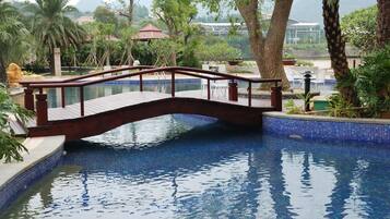 Un puente de madera sobre una piscina azul cristalina con bordes de azulejos, rodeado de exuberante follaje y árboles.