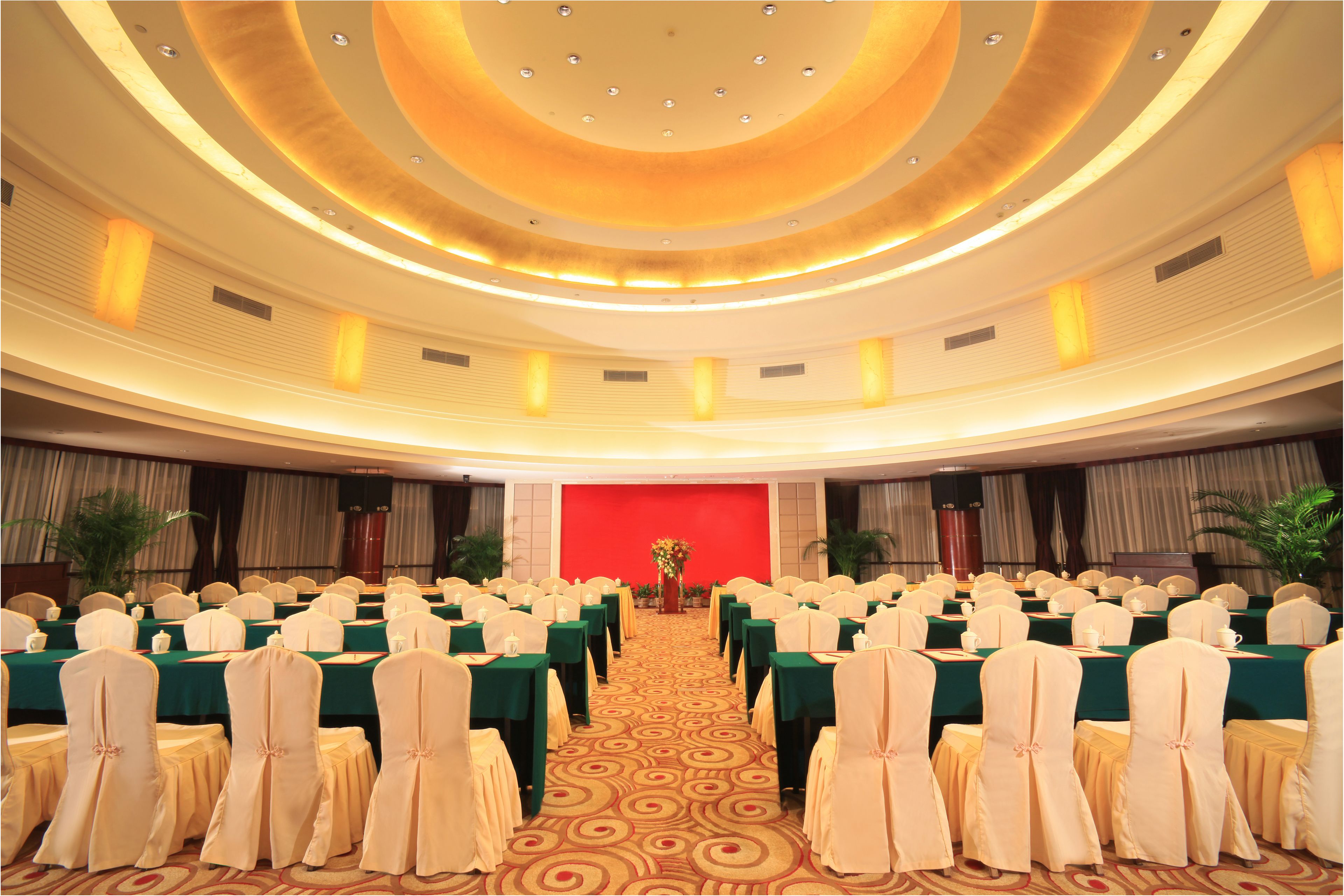 A banquet hall with rows of tables and chairs, a central stage, and a red backdrop.