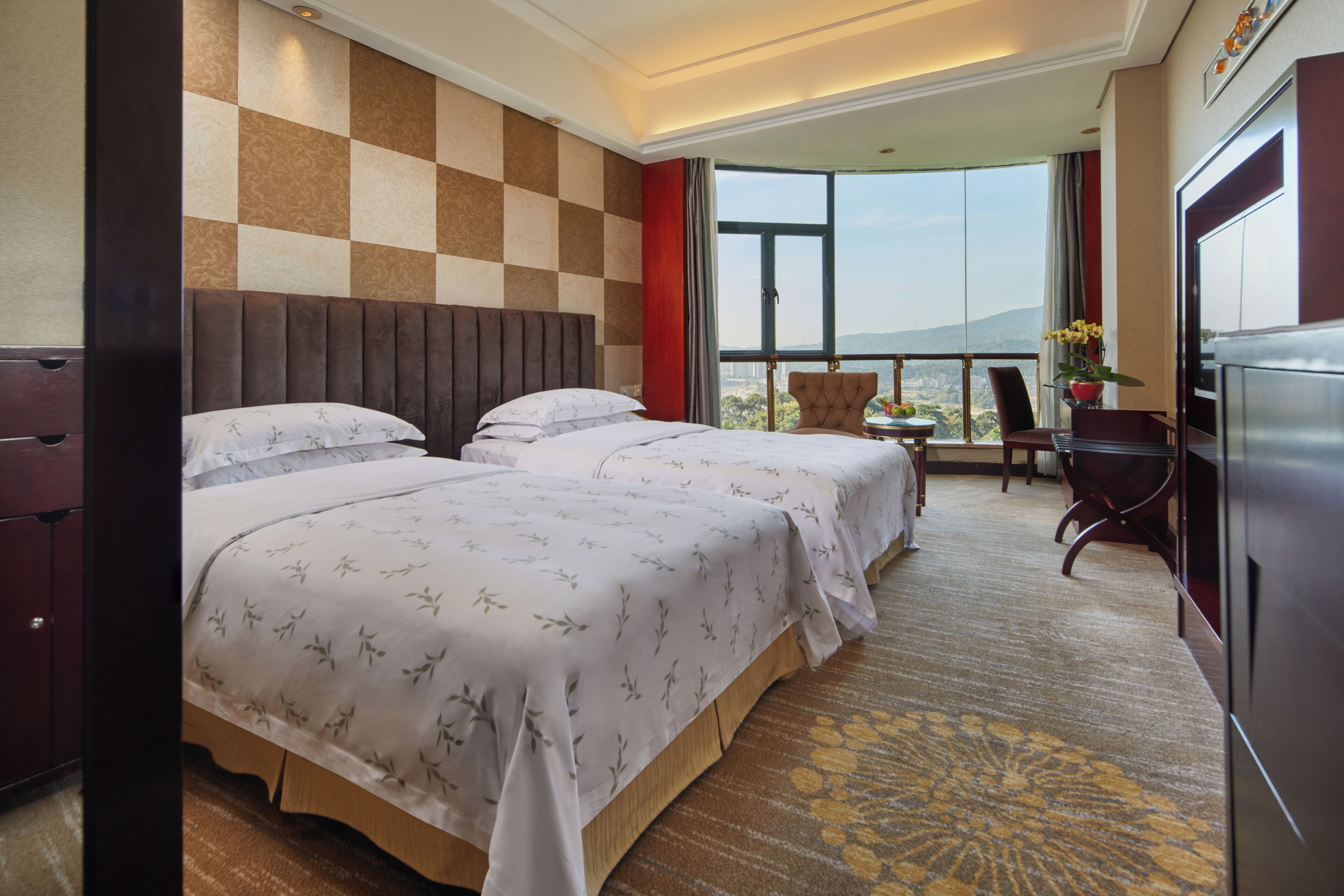 A hotel room with two beds, a large window with a city view, a dining area with a table and chairs, and a TV.