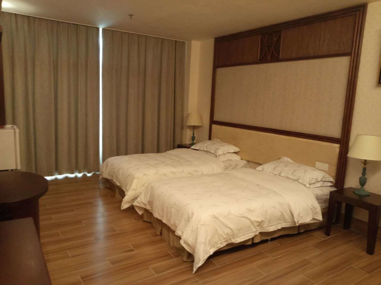 Habitación de hotel con dos camas, piso de madera, un ventanal grande con cortinas y una mesita con una lámpara.