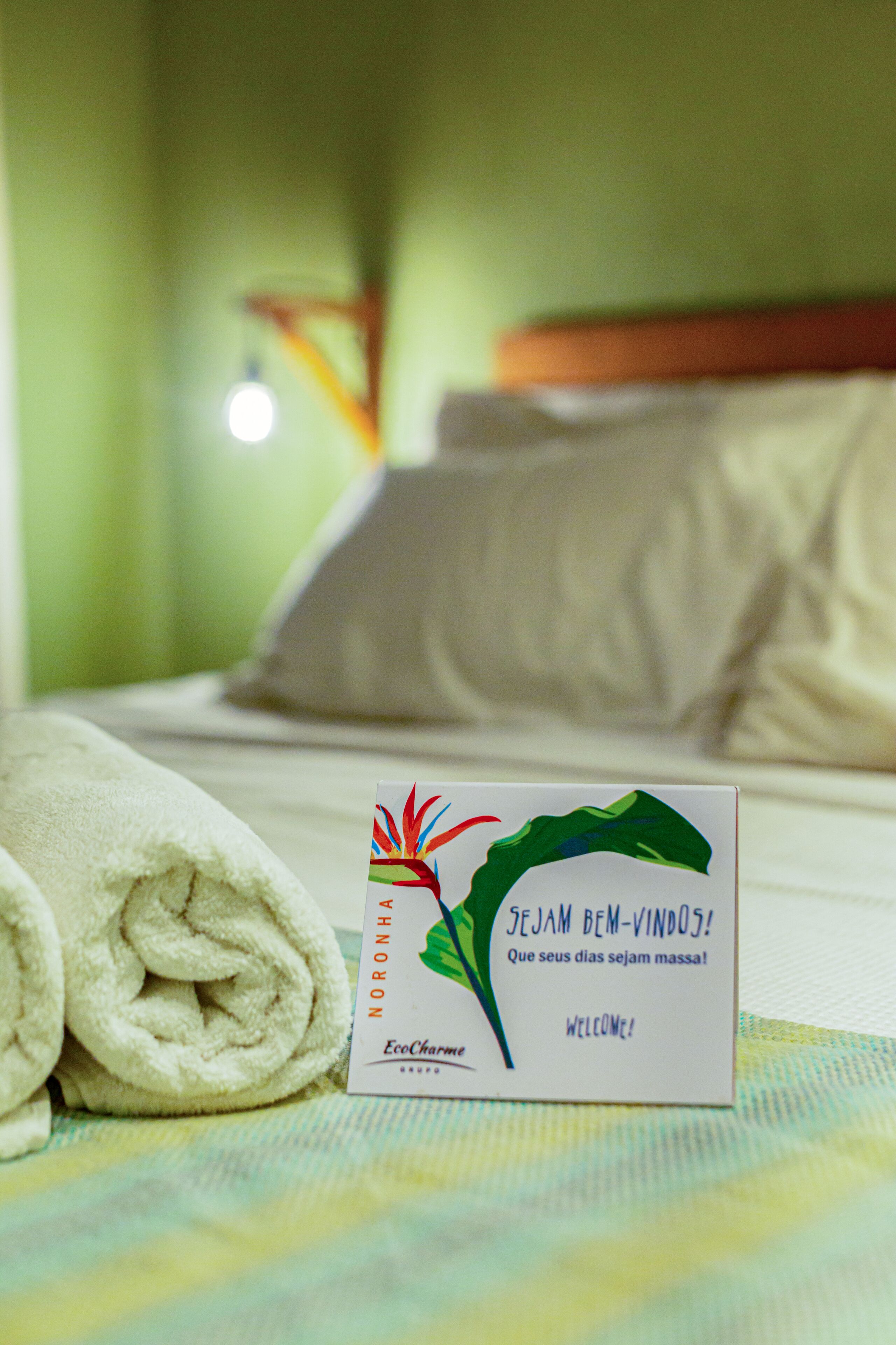 A neatly made bed with a rolled white towel and a welcome card on a bedspread.