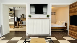 A modern living room with a flat-screen TV mounted above a white radiator, a sofa with cushions, and a dining area with a wooden table and chairs. - Hotel Nanis (Wunstorf)