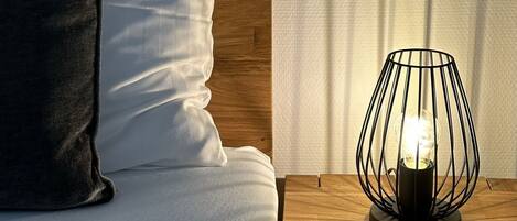 Una cama bien hecha con un almohadón blanco y uno oscuro, una mesita de noche de madera con una lámpara moderna, y un cabecero de madera.