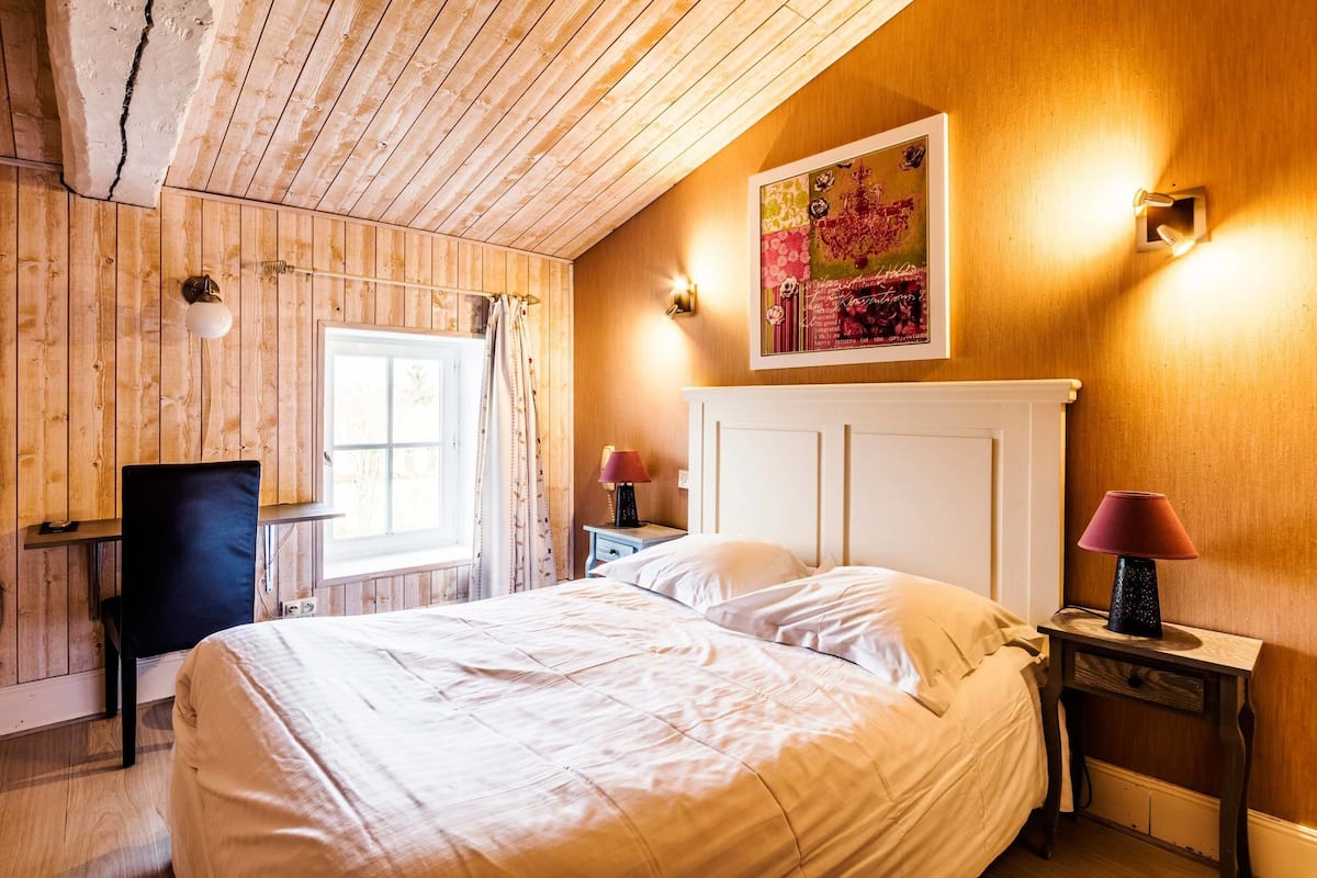 Un dormitorio con techo de madera, una cama con ropa de cama blanca, un escritorio con una silla y una ventana con cortinas.
