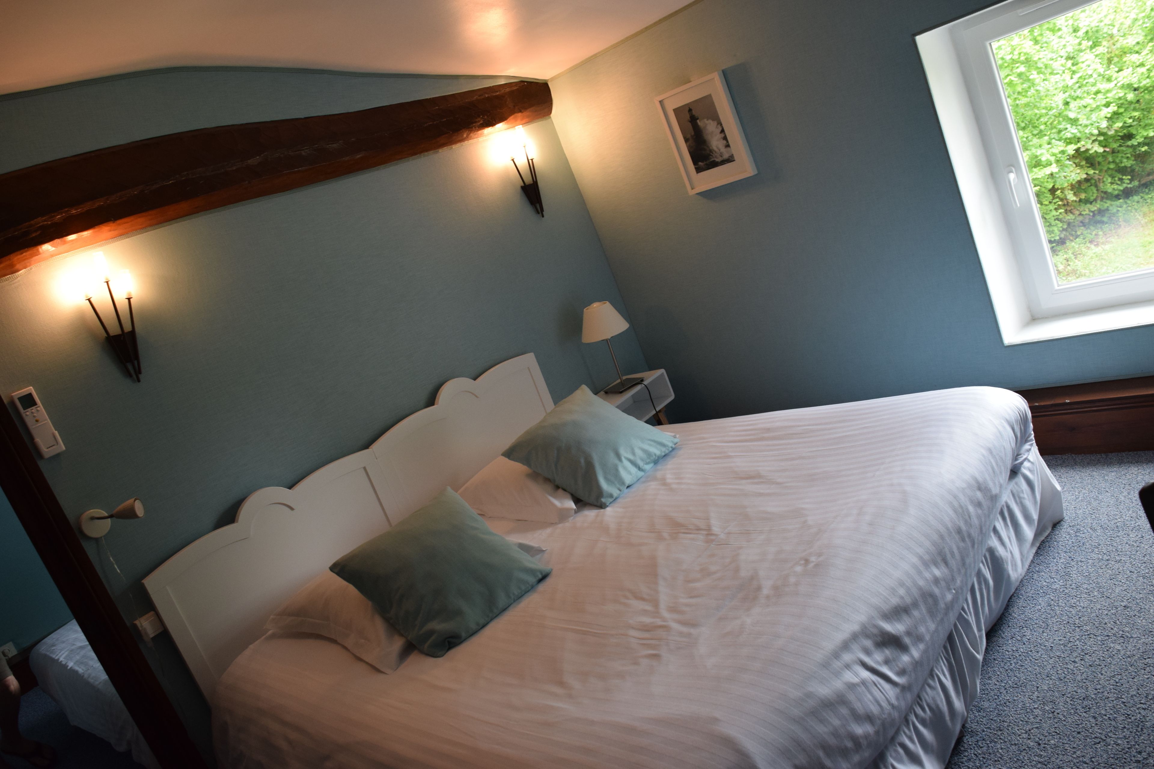 A bedroom with a bed, pillows, a nightstand, a lamp, a wall-mounted light fixture, a window, and a framed picture on the wall.