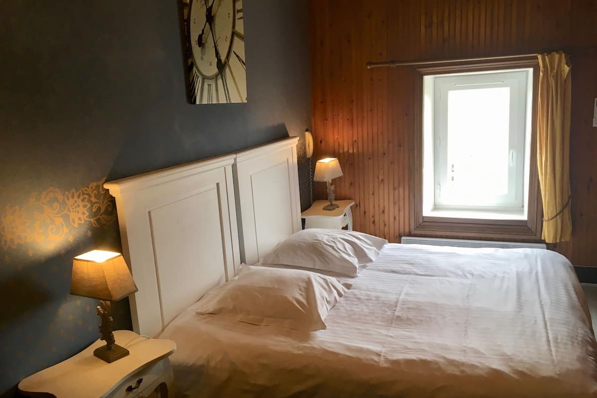 Un dormitorio con una cama, dos mesitas de noche, un reloj de pared y una ventana con cortinas.