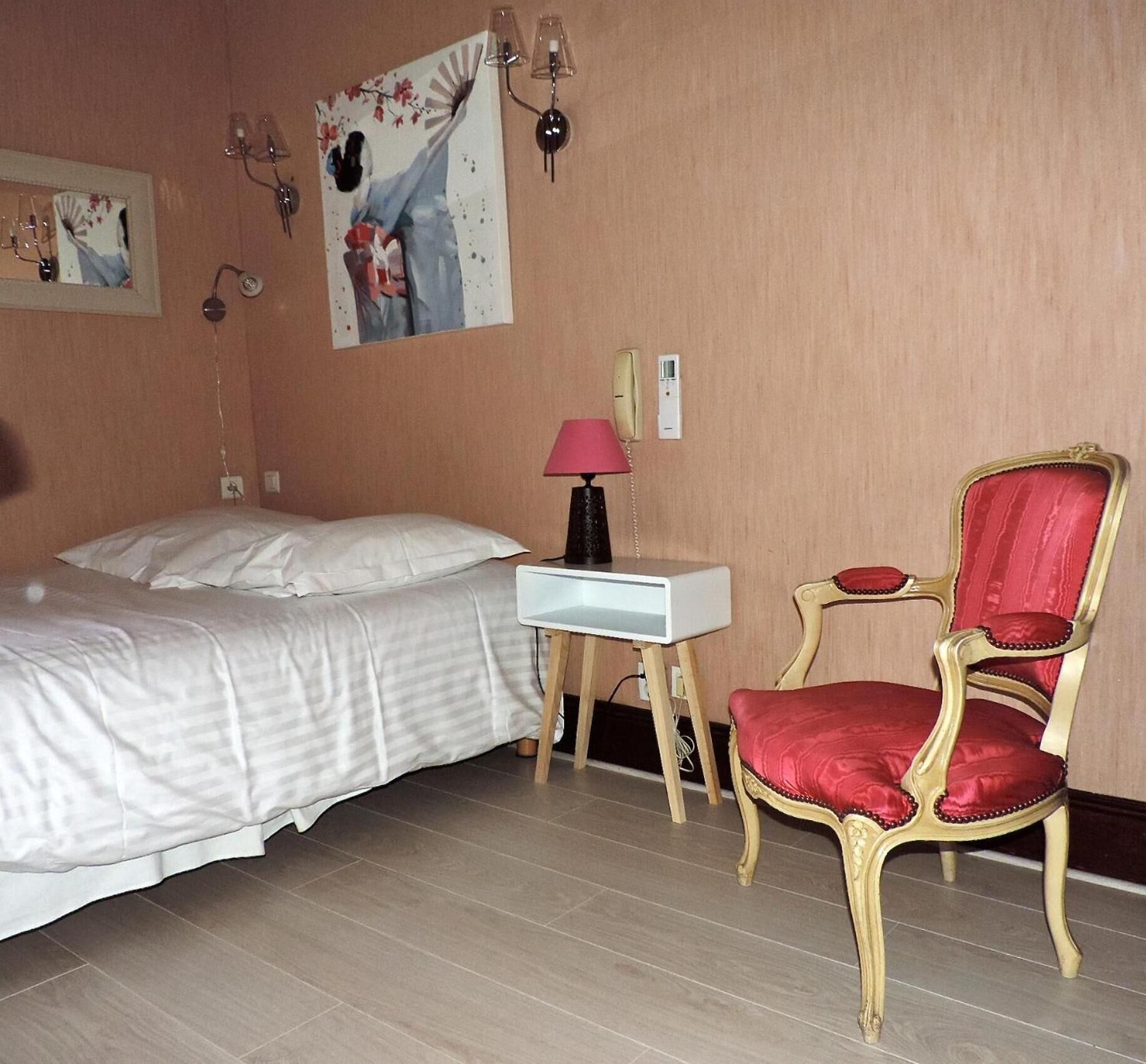 A hotel room with a bed, a desk with a lamp, a chair, and two paintings on the wall.