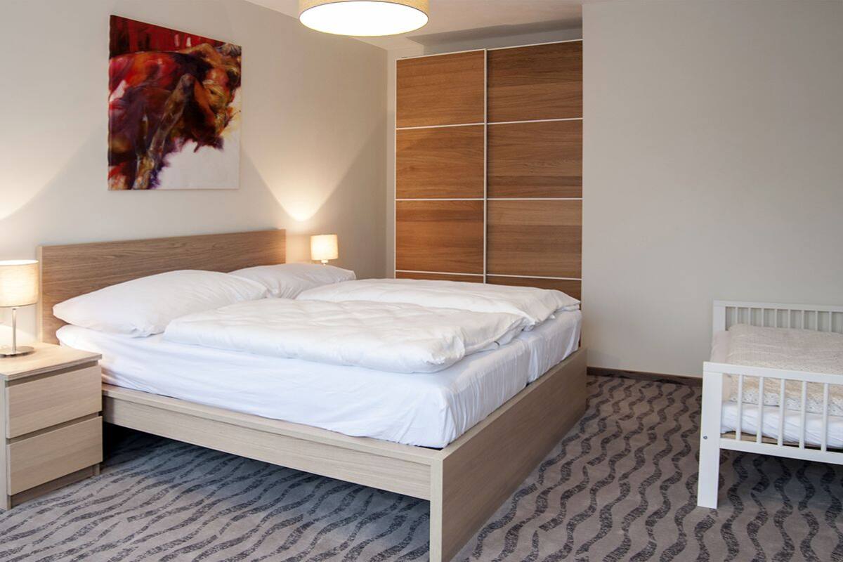 A bedroom with a wooden bed, a nightstand with a lamp, a crib, and a large wooden wardrobe.