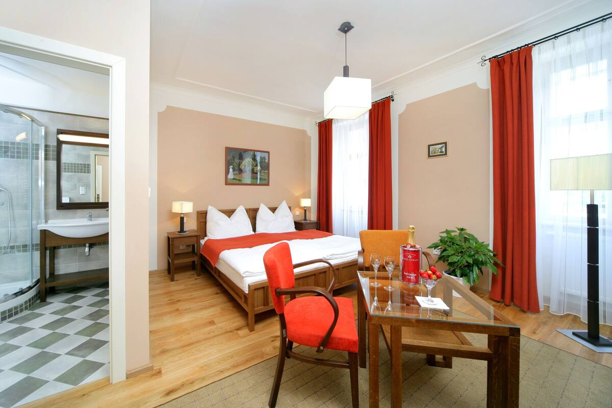 A hotel room with a bed, bedside tables, a desk with chairs, a glass shower enclosure, and a potted plant.