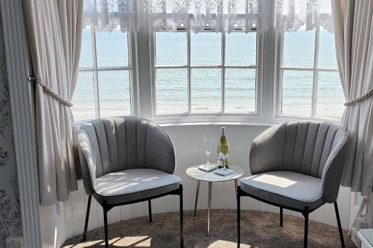 Ein Zimmer mit zwei grauen Polstersesseln, die zu einem Fenster mit Meerblick ausgerichtet sind. Ein kleiner Tisch mit Weinflasche und Glas steht zwischen den Sesseln.