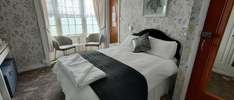 A hotel room with a bed, a desk, a chair, a window with a view of the sea, and floral wallpaper.