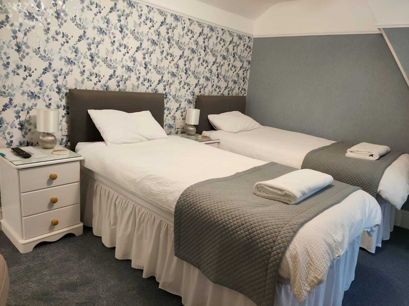 A hotel room with two beds, a nightstand with a lamp, and a floral wallpaper.
