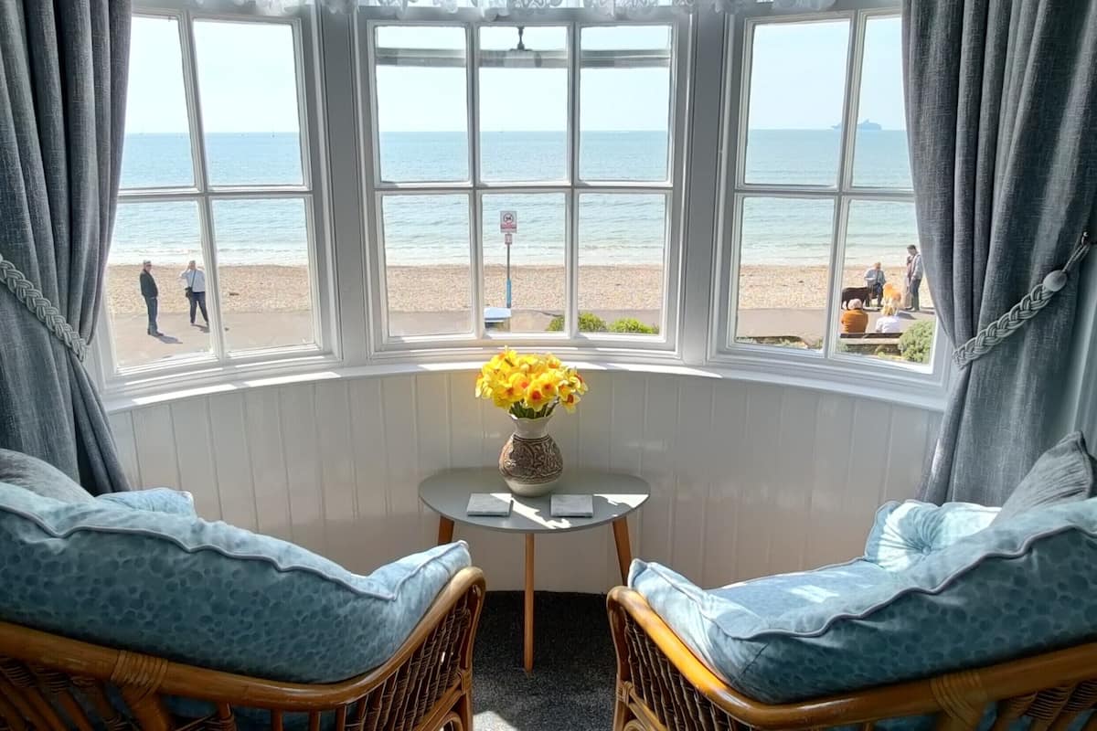 Ein Zimmer mit Erkerfenster und Blick auf einen Strand, mit Korbmöbeln, einem kleinen Tisch mit Blumenvase und Menschen, die am Strand sind.
