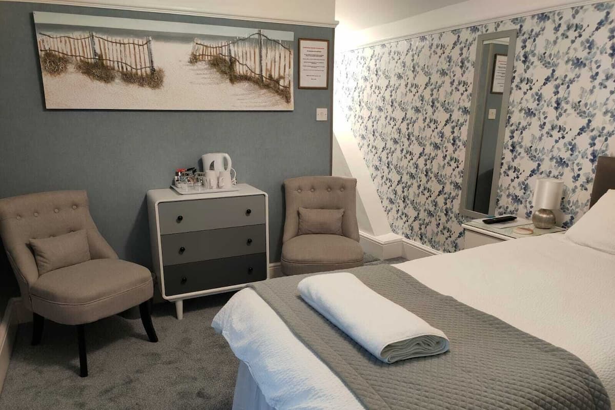 Ein Hotelzimmer mit einem Bett, zwei Sesseln, einem Schminktisch, einem Gemälde und blumigem Tapetenmuster.
