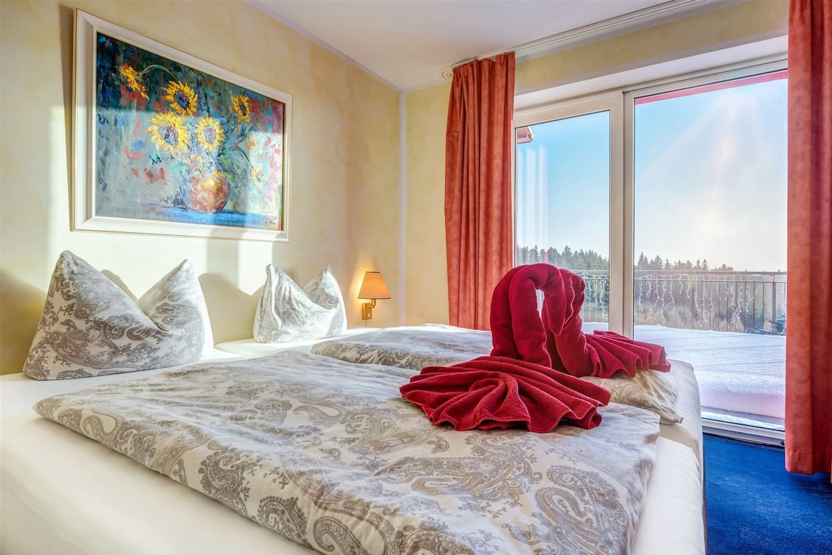 Ein Hotelzimmer mit einem großen Bett, einem Gemälde an der Wand, einem Fenster mit Blick auf Bäume und einem roten Handtuch auf dem Bett.