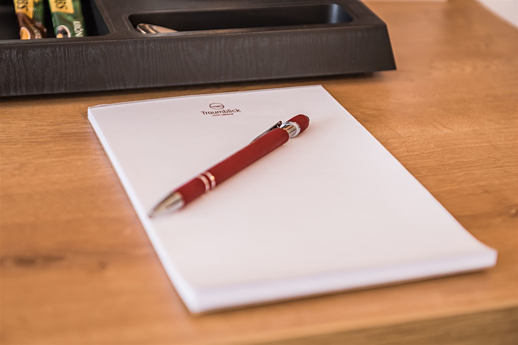 A white notepad with a red pen on a wooden table, a black tray with pens, and a bottle of hand sanitizer.