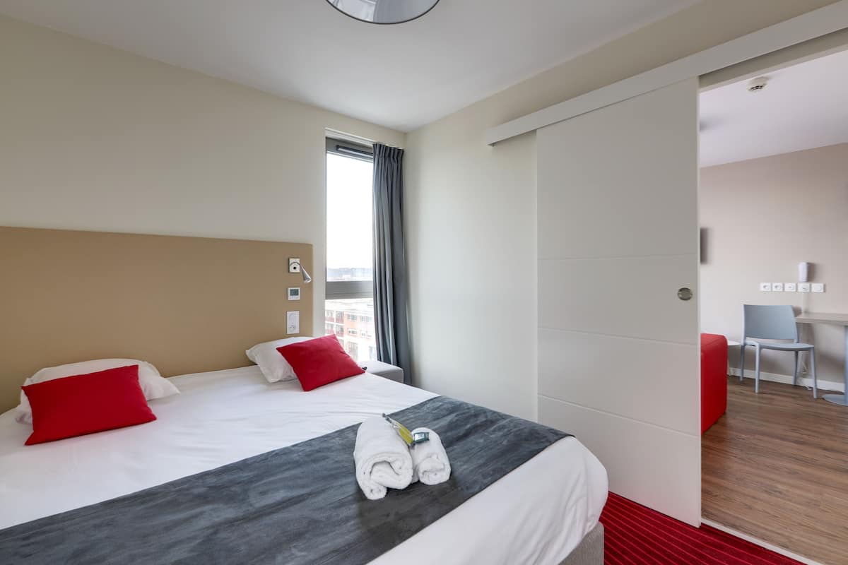 Une chambre d’hôtel avec un lit, deux oreillers rouges, une tête de lit grise et une fenêtre avec des rideaux.