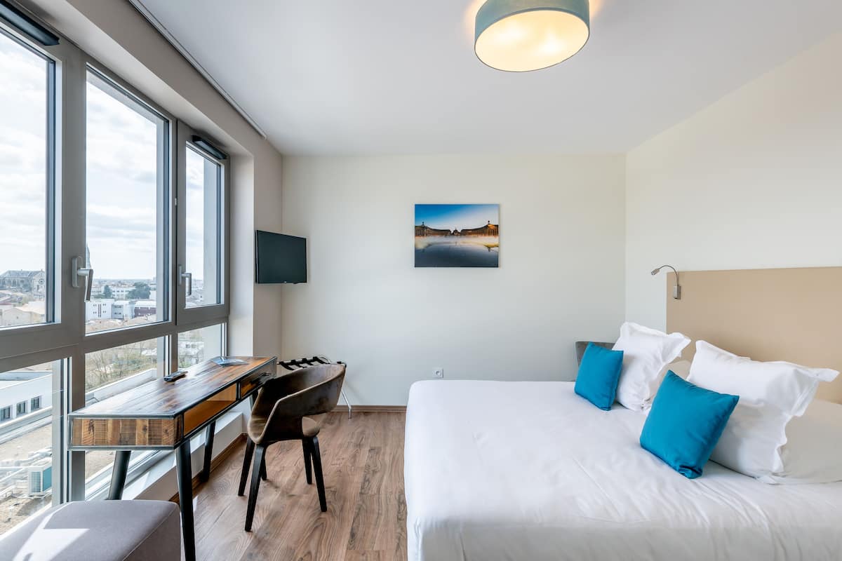 Une chambre d’hôtel moderne avec une grande fenêtre, un bureau, un lit avec des oreillers bleus et un tableau au mur.