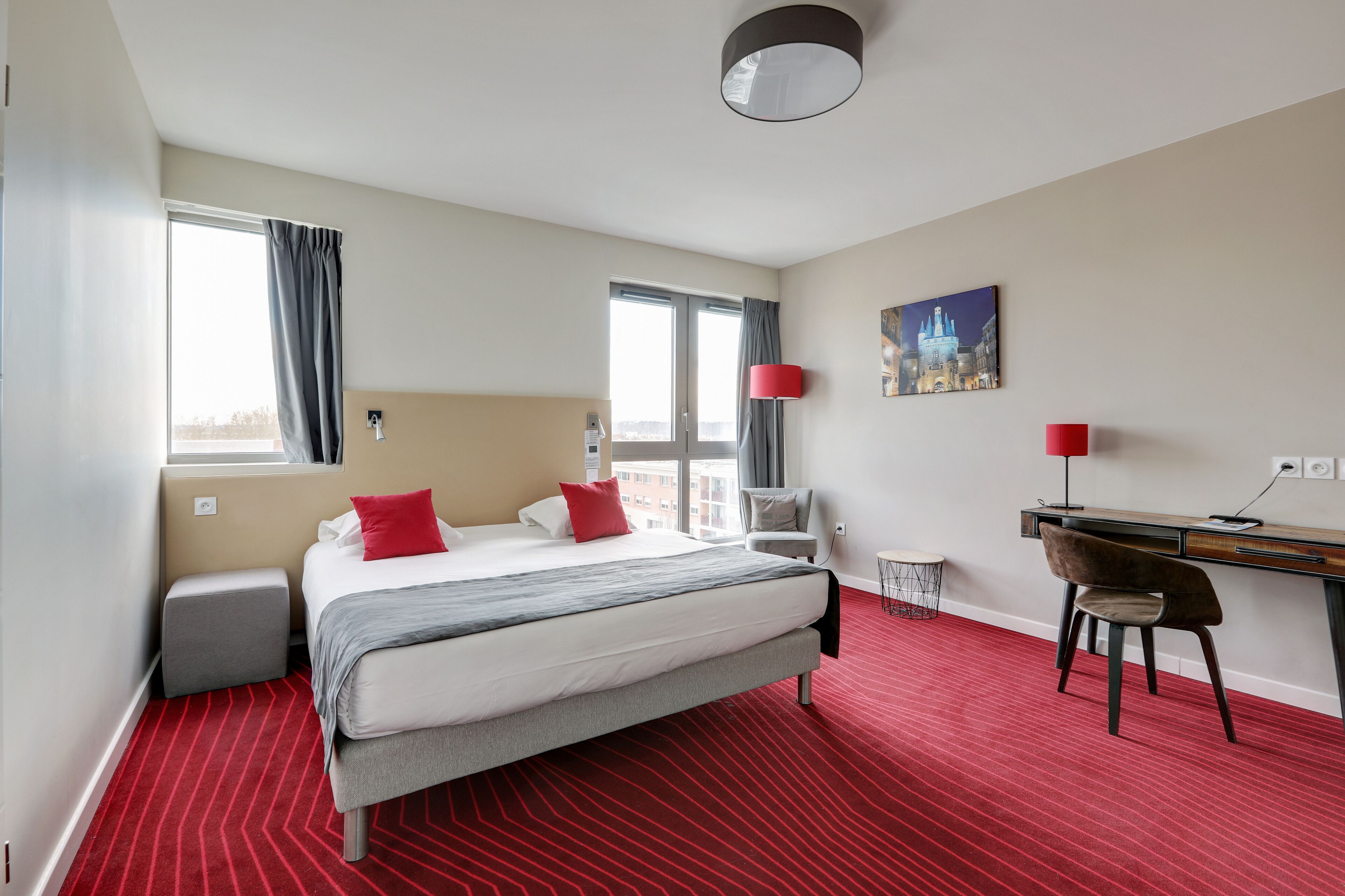 A hotel room with a bed, a desk, a chair, a lamp, and a painting on the wall.